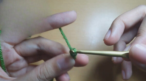 Green yarn looped around fingers as a slip knot tightens on the hook.