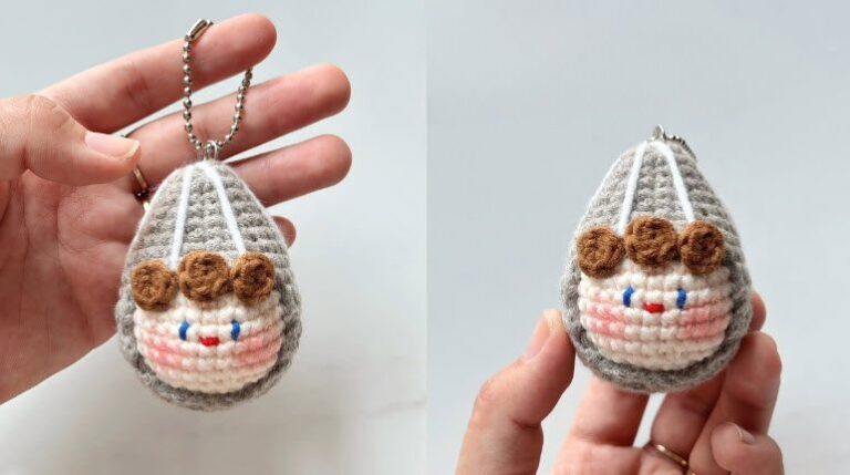 Gray egg-shaped crochet keychain with a smiling cream face, brown curl bangs and pink blush, held in hand